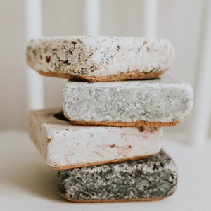 May include: A stack of four square-shaped coasters made of natural stone. The coasters are in various shades of gray, white, and brown. The coasters have a rough, textured surface.