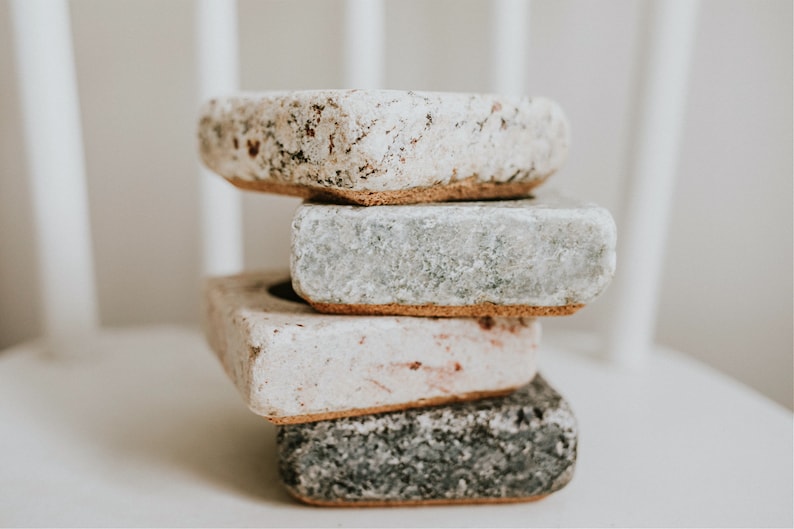 May include: A stack of four square-shaped coasters made of natural stone. The coasters are in various shades of gray, white, and brown. The coasters have a rough, textured surface.