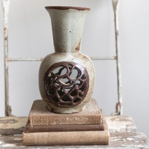 May include: A white ceramic vase with a brown cutout design. The vase is sitting on a stack of three books.
