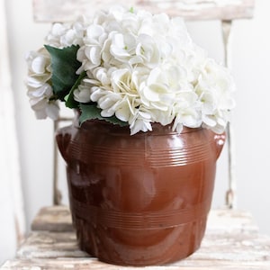 May include: A brown ceramic vase with a glossy finish, filled with white hydrangea flowers and green leaves. The vase has a ribbed design and two small handles. The arrangement sits on a distressed wooden chair.