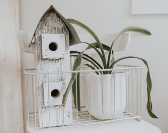 May include: A white birdhouse with two openings sits in a white wire basket with a potted green plant. The birdhouse has a weathered wood look and the plant has long, thin leaves.