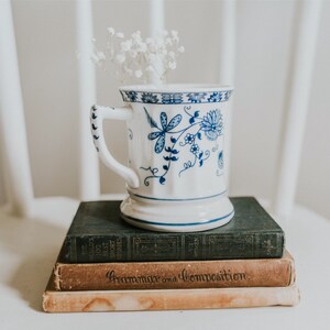 Puede incluir: Una taza de cer&aacute;mica blanca con un dise&ntilde;o floral azul, llena de flores blancas, se encuentra sobre una pila de tres libros. El libro superior es de color verde oscuro, el libro del medio es marr&oacute;n con el texto "Grammar and Composition" impreso en el lomo, y el libro inferior es de color marr&oacute;n claro.