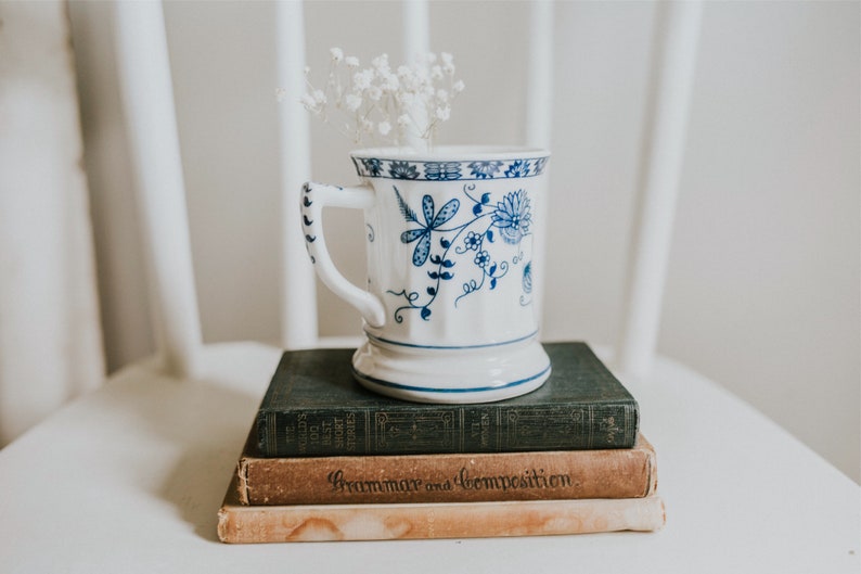 Puede incluir: Una taza de cer&aacute;mica blanca con un dise&ntilde;o floral azul, llena de flores blancas, se encuentra sobre una pila de tres libros. El libro superior es de color verde oscuro, el libro del medio es marr&oacute;n con el texto "Grammar and Composition" impreso en el lomo, y el libro inferior es de color marr&oacute;n claro.