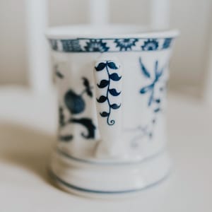 Puede incluir: Una taza de cer&aacute;mica blanca con un dise&ntilde;o floral azul y blanco. El asa de la taza tiene un dise&ntilde;o de bigote azul y blanco.
