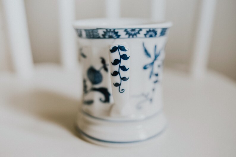 Puede incluir: Una taza de cer&aacute;mica blanca con un dise&ntilde;o floral azul y blanco. El asa de la taza tiene un dise&ntilde;o de bigote azul y blanco.