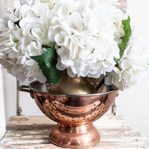 May include: A copper colander with a white hydrangea bouquet inside. The colander has a stand and is sitting on a whitewashed wooden surface.