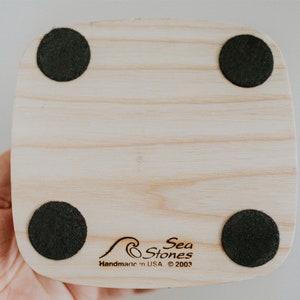 May include: The bottom of a wooden coaster with four black felt pads. The coaster has the text "Sea Stones" and "Handmade in USA &copy; 2003" engraved on it.