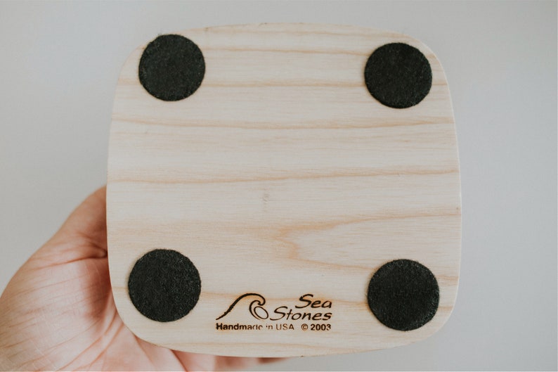 May include: The bottom of a wooden coaster with four black felt pads. The coaster has the text "Sea Stones" and "Handmade in USA &copy; 2003" engraved on it.