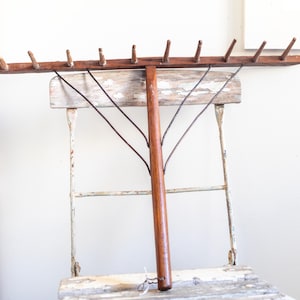 May include: A vintage wooden rake with a long, dark brown handle and a horizontal head with several wooden pegs. The rake is resting on a distressed, white-painted chair, creating a rustic aesthetic.