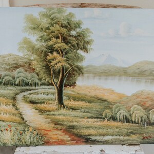 Op de afbeelding: Een schilderij van een landschap met een kronkelend pad dat naar een meer leidt. Een grote boom staat op de voorgrond, met bergen in de verte. De lucht is lichtblauw met witte wolken.