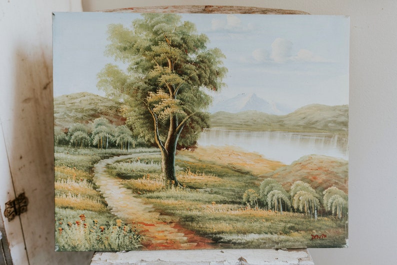 Op de afbeelding: Een schilderij van een landschap met een kronkelend pad dat naar een meer leidt. Een grote boom staat op de voorgrond, met bergen in de verte. De lucht is lichtblauw met witte wolken.