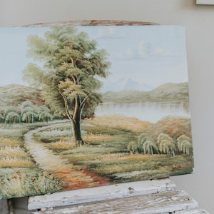 Op de afbeelding: Een vintage olieverfschilderij dat een landschapsscene voorstelt met een kronkelend pad dat naar een meer leidt, omringd door heuvels en bomen. Het schilderij toont een grote boom op de voorgrond en een bergketen op de achtergrond.