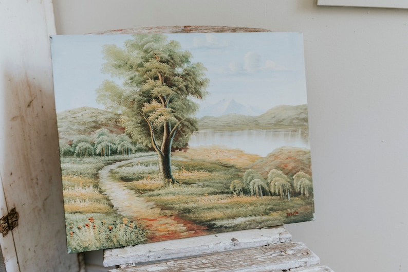 Op de afbeelding: Een vintage olieverfschilderij dat een landschapsscene voorstelt met een kronkelend pad dat naar een meer leidt, omringd door heuvels en bomen. Het schilderij toont een grote boom op de voorgrond en een bergketen op de achtergrond.