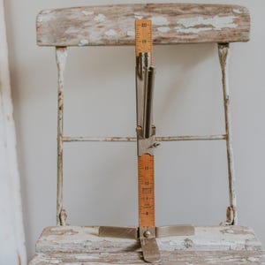 May include: A vintage wooden ruler with metal arms and a base, standing on a white painted chair. The ruler has markings in inches from 1 to 20.
