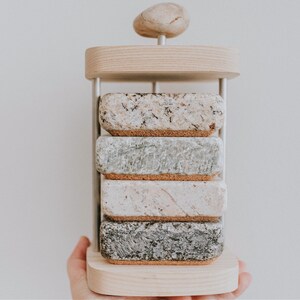 May include: A set of four stone coasters with cork backing, stacked on a wooden stand with a round stone on top. The coasters are in various shades of gray, white, and brown.