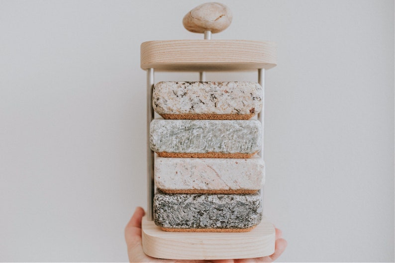 May include: A set of four stone coasters with cork backing, stacked on a wooden stand with a round stone on top. The coasters are in various shades of gray, white, and brown.