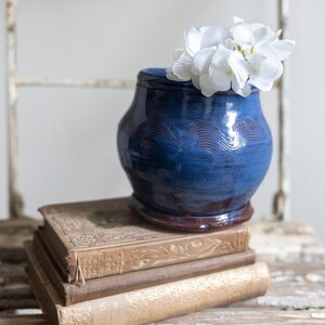 May include: A blue ceramic vase with a wavy design sits on top of a stack of old books. White flowers are in the vase.