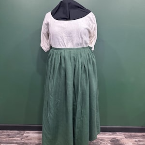 Puede incluir: Un vestido de estilo histórico sobre un maniquí. El vestido presenta una parte superior blanca de manga corta y una falda larga verde plisada. Una capucha negra cubre la cabeza del maniquí. El vestido se exhibe contra una pared verde.