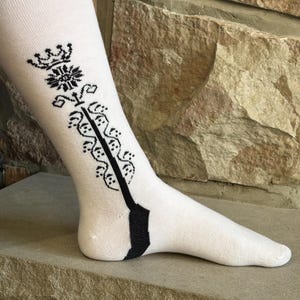 May include: White sock with a black decorative design on the calf and heel. The design includes a crown, floral elements, and a vertical stripe. The sock is displayed on a stone surface against a stone wall.