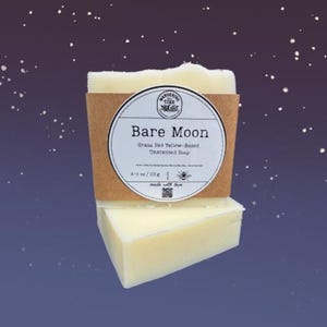 以下が含まれることがあります： 2つの無香料石鹸で、1つはもう1つの上に積み重ねられています。上の石鹸は「Bare Moon」の文字とその他のテキストが書かれた茶色の紙ラベルで包まれています。石鹸は淡い黄色です。