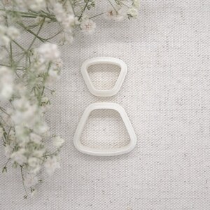 May include: Two white plastic cookie cutters shaped like a dress. The top cutter is a small, rounded triangle shape. The bottom cutter is a larger, rounded triangle shape.