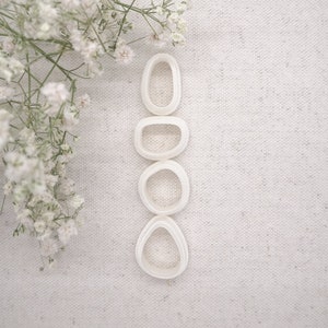 May include: Four white polymer clay cutters in various organic shapes, including an oval, a rounded rectangle, a circle, and a teardrop. The cutters are arranged vertically on a textured, off-white surface, with delicate white flowers in the upper left corner.