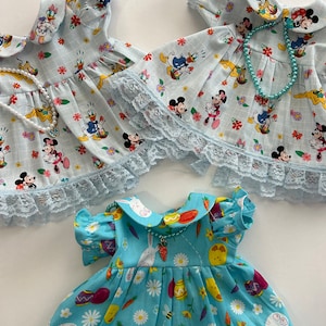 Op de afbeelding: Drie blauwe en witte poppenjurken met kantversiering. De eerste twee jurken hebben een Mickey Mouse en Donald Duck print. De derde jurk heeft een konijn, kuiken en wortel print.