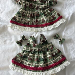 May include: A set of two green dresses featuring a teddy bear and white picket fence print, accented with dark pink trim and white eyelet lace ruffles. The set includes a pair of white bloomers and two matching hair bows with the same bear print.