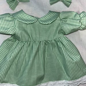 Puede incluir: Un vestido de cuadros verdes y blancos con un ribete de encaje blanco, junto con dos lazos para el pelo a juego. El vestido presenta un cuello Peter Pan y mangas abullonadas, perfecto para un bebé o niño pequeño.