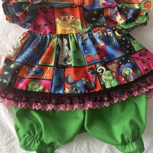May include: A two-piece outfit consisting of a short-sleeved top and green bloomers. The top features a colorful Halloween-themed print with pumpkins, bats, and cartoon characters, accented by a black lace trim and a small orange pumpkin button.