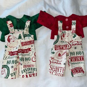 Könnte beinhalten: Zwei Puppenkleidungssets bestehend aus einem langärmeligen Hemd und einer Latzhose. Ein Set enthält ein dunkelgrünes Hemd und eine cremefarbene Latzhose mit Aufdrucken von Weihnachtsmann, Tannenbäumen und Texten wie "Winter Wonderland" und "Ho Ho Ho".