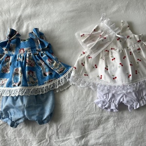 Puede incluir: Dos conjuntos para muñeca compuestos por tops sin mangas y bombachos. El conjunto izquierdo es azul con estampado de personajes y encaje blanco. El conjunto derecho es blanco con estampado de cerezas rojas, encaje y un lazo para el pelo a juego.