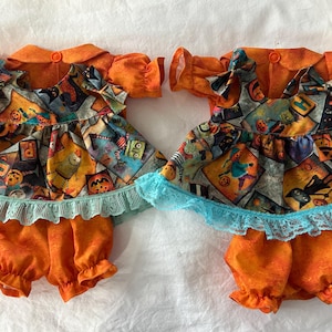 May include: Two matching doll outfits featuring orange textured fabric shirts and bloomers. The tops have a multi-coloured Halloween print with pumpkins, cats, and monsters, finished with a light blue lace trim along the bottom hem.