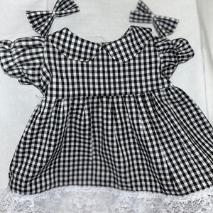 May include: A black and white gingham baby dress with a Peter Pan collar, short puffed sleeves, and a white lace hem. Two matching gingham bows are placed above the dress.