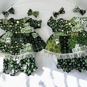 May include: Two St. Patrick's Day outfits with matching bows. The dresses feature a patchwork design with green and black clover patterns, plaid, and the word "Lucky". Each outfit includes a dress and bloomers, trimmed with white lace.