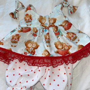 May include: A two-piece baby outfit featuring a light blue top with a repeating pattern of teddy bears and red accents. The top has red lace trim and bows. The white pants have a red heart pattern and ruffled hems.