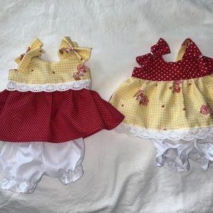 May include: Two baby outfits. One outfit has a yellow gingham top with red polka dot skirt and white bloomers. The other outfit has a red polka dot top with yellow gingham skirt and white bloomers. Both outfits have white lace trim.