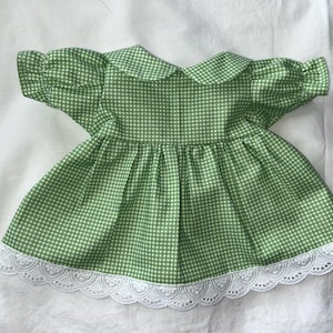 Puede incluir: Un vestido de cuadros vichy verdes y blancos con mangas cortas abullonadas, cuello redondo y falda fruncida. El dobladillo del vestido está rematado con una ancha banda de encaje blanco festoneado, que aporta un borde decorativo.