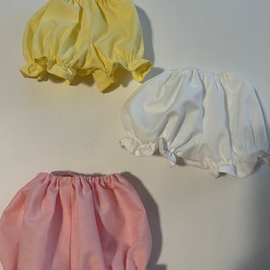 May include: Three pairs of baby bloomers in yellow, white, and pink. The bloomers have a gathered elastic waistband and a ruffled hem.