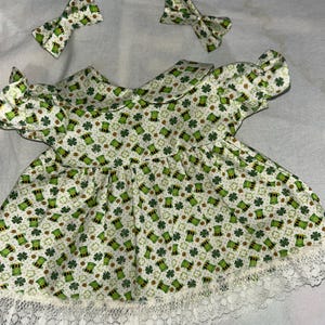 May include: A white dress with a green and gold print of shamrocks and leprechaun hats. The dress has a white lace trim and comes with two green bows.