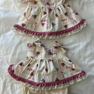 May include: A two-piece doll outfit featuring a cream-colored top and matching bloomers. The top has short sleeves, a pink button, and a print of cakes and party text. The hem is finished with a dark pink ruffle and white lace trim.
