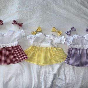 Peut inclure: Trois robes de poupée avec des nœuds pour les cheveux assortis. Chaque robe a un haut blanc avec un col Claudine et une bordure en dentelle, et une jupe à carreaux vichy rouge, jaune et violet. Les nœuds sont assortis aux couleurs des jupes.