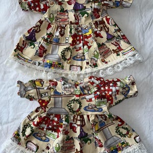 Może przedstawiać: Dwie kremowe sukienki dla niemowląt z nadrukiem o tematyce bożonarodzeniowej z elfami, Świętym Mikołajem i prezentami. Sukienki mają krótkie rękawy, kołnierzyk Piotrusia Pana i białe koronkowe wykończenie wzdłuż dołu. Tkanina zawiera tekst, taki jak "Apples" i "Santa's Chair".