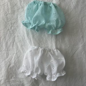 Może przedstawiać: Dwie pary bloomers z falbankami. Jedna para jest jasnoniebieska z haftowanymi kwiatowymi detalami. Druga para jest biała z podobnym haftem kwiatowym. Obie mają elastyczne paski w talii i falbaniaste obszycia, wykonane z lekkiej tkaniny.