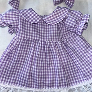 以下が含まれることがあります： 白と紫のギンガムチェックのドレスで、白いレースのトリムとそれに合わせたリボンが付いています。ドレスはピーターパンカラーと短い袖です。