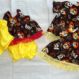 Puede incluir: Dos vestidos de muñeca con temática de Acción de Gracias. Un vestido es marrón con un ribete de encaje amarillo y el otro es marrón con un ribete de encaje rojo. Ambos vestidos tienen un patrón de pavos y calabazas.