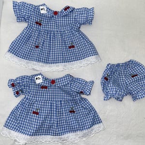 May include: Two blue and white gingham dresses with red cherry embroidery and white lace trim, plus matching bloomers. The dresses have short sleeves and red button details. The dresses and bloomers are in a classic style.