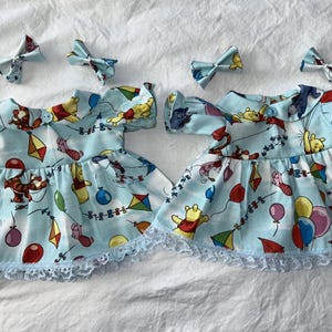 Op de afbeelding: Twee lichtblauwe babyjurkjes met bijpassende haarstrikken. De jurkjes hebben een Winnie de Poeh-print met ballonnen, vliegers en personages. Elk jurkje heeft een witte kanten rand aan de zoom en korte mouwen. De strikken hebben dezelfde print.