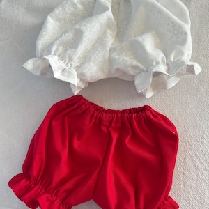 Può includere: Un top bianco con motivo a fiocchi di neve e bloomers rossi. Il top ha maniche a balze e scollatura arricciata. I bloomers sono di colore rosso pieno con orli a balze. Il set è realizzato in tessuto morbido.
