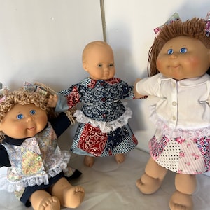 May include: Three Cabbage Patch Kids dolls wearing different patchwork outfits. The dolls have blonde hair and blue eyes. One doll is wearing a blue and red patchwork dress with a white lace trim. Another doll is wearing a white shirt with a white lace trim and a pink and white patchwork skirt. The third doll is wearing a blue and white patchwork dress with a white lace trim.